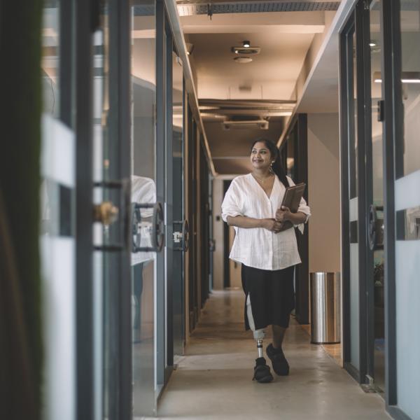 Woman worker with prosthetic limb holding her digital tablet and walking in an office corridor smiling